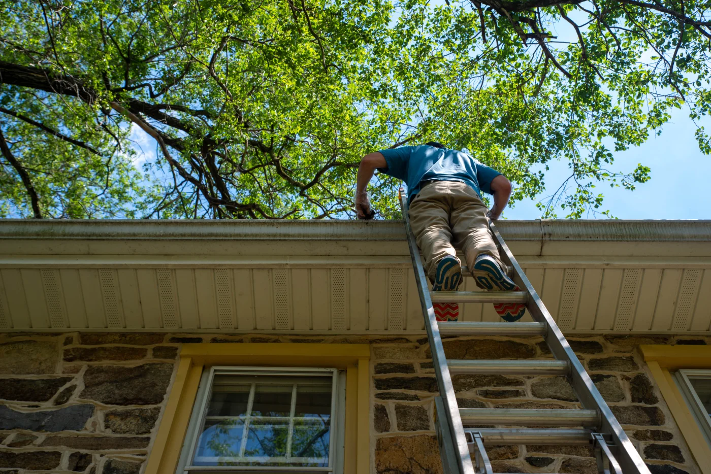 An upward view of a roofer climbing onto a roof to make repairs on a stone house after storm damage.