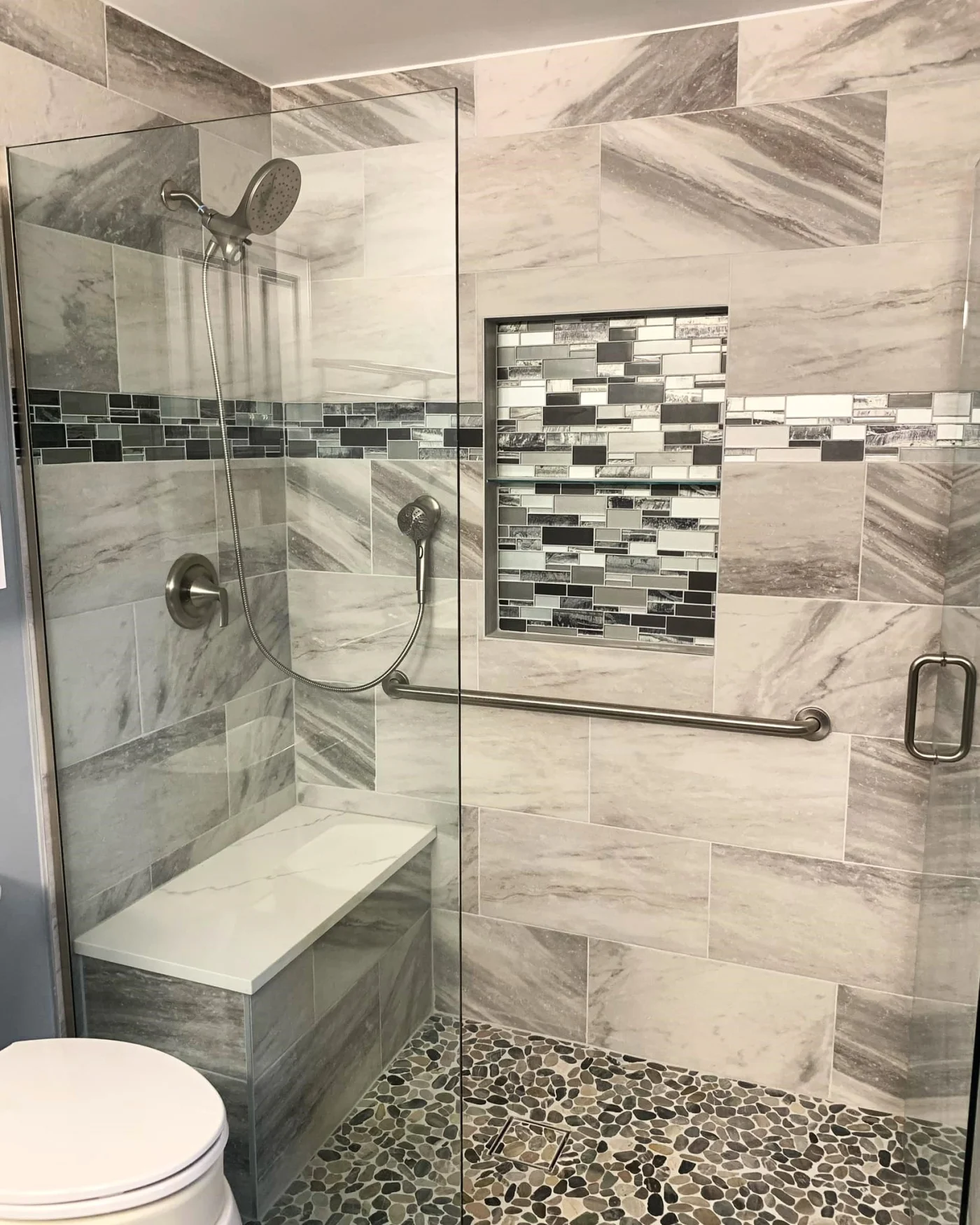 A tan walk-in shower with a glass door, a bench, built-in shelves, a hand rail, and silver fixtures.