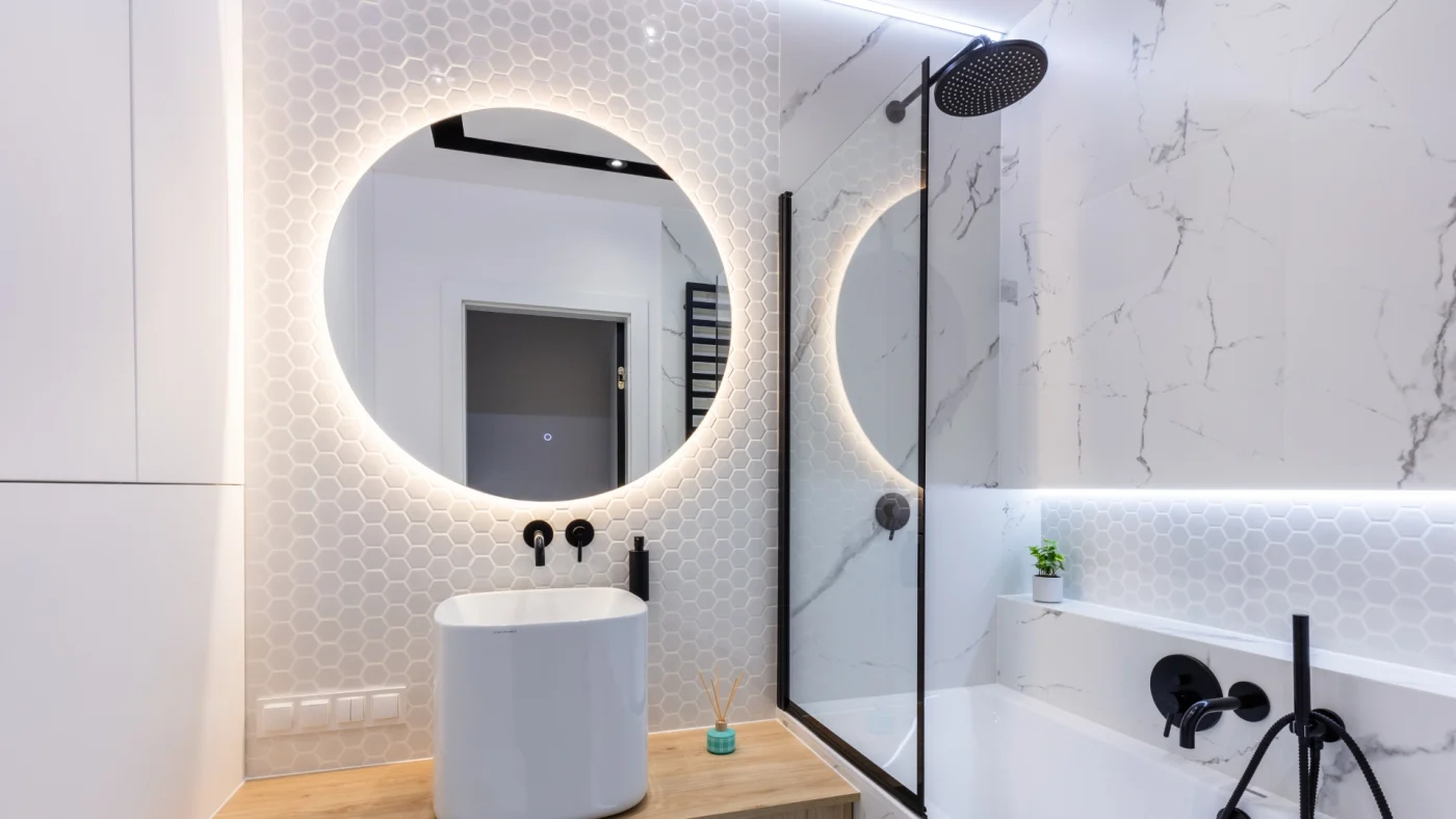 A white bathroom with a wooden vanity, a white sink, a circular mirror on the wall, and a white bathing space.