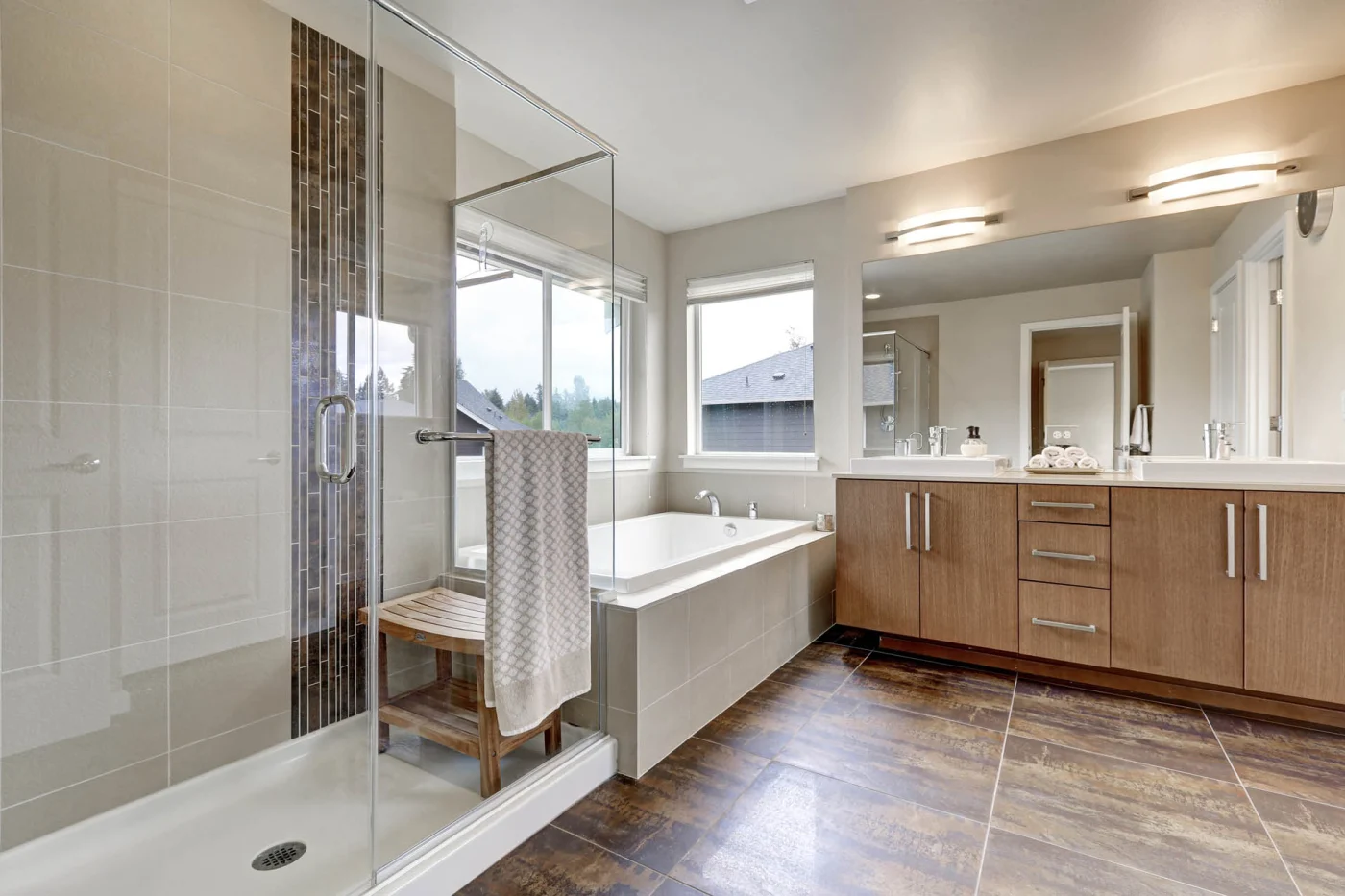 A white bathroom with a wooden vanity, a large mirror with overhead lighting, a glass walk-in shower, and a white bathtub with three windows next to it.